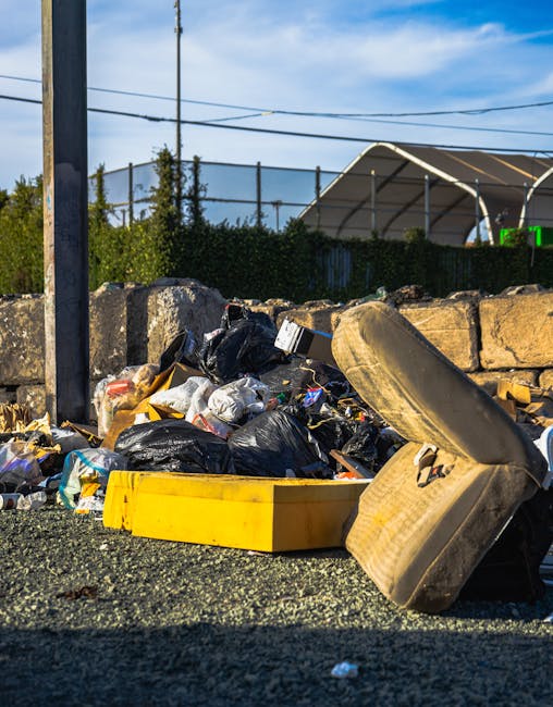 A collection of assorted household rubbish and waste materials piled on the ground in an outdoor area, including black plastic trash bags, a yellow plastic container, and a discarded car tire leaning 